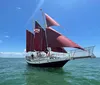 A sailboat with red sails is navigating the sea under a clear blue sky with several people visible on deck