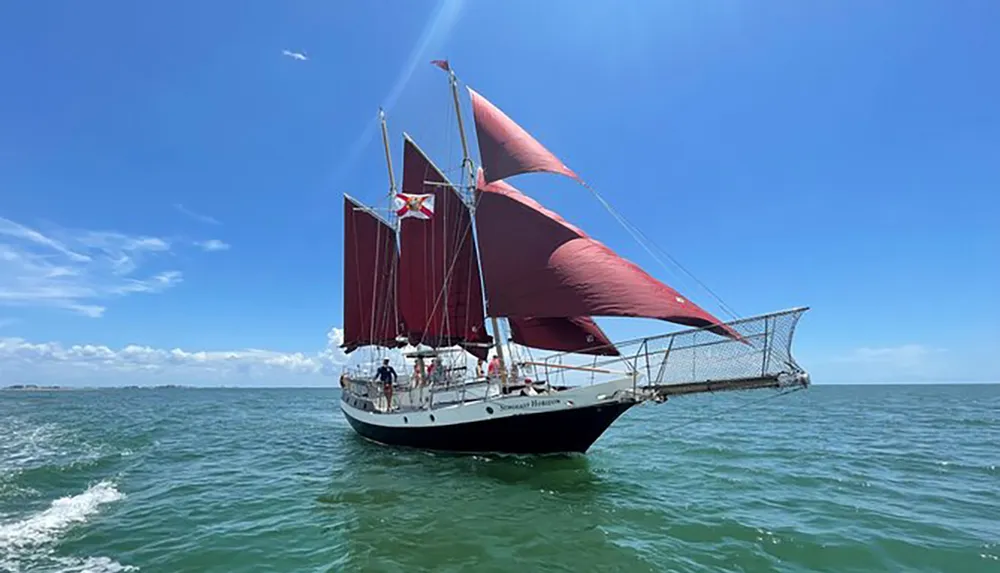 A sailboat with red sails is navigating the sea under a clear blue sky with several people visible on deck