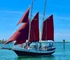 A sailboat with red sails is navigating the sea under a clear blue sky with several people visible on deck