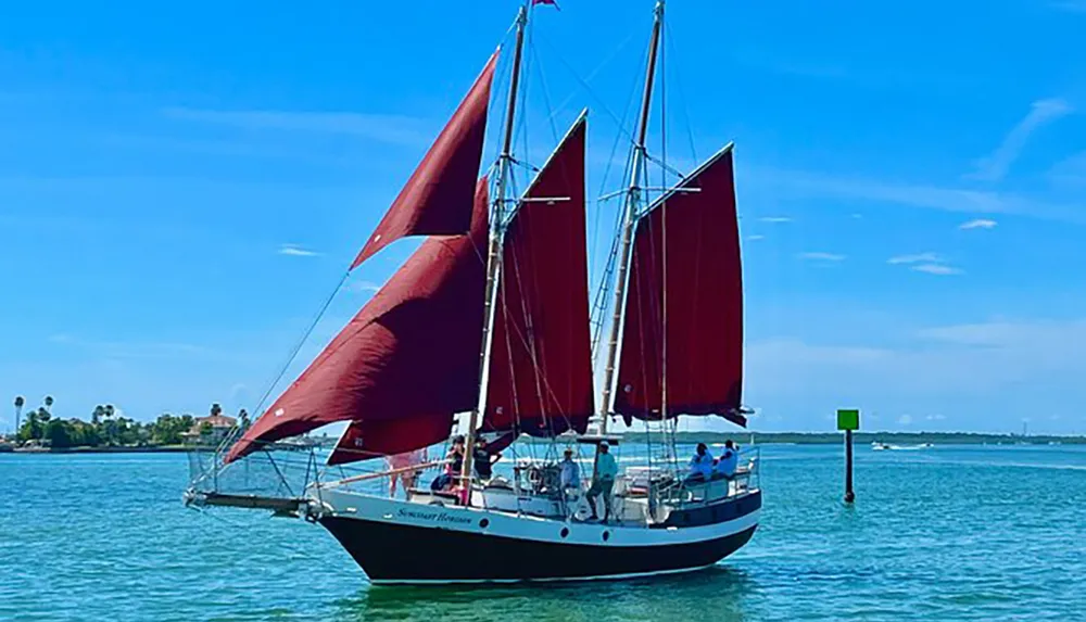 A schooner with striking red sails cruises through calm blue waters under a clear sky