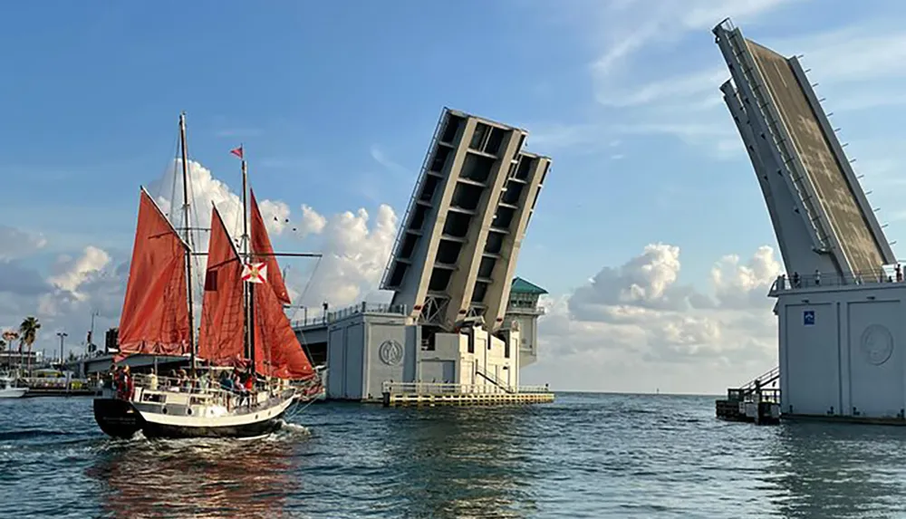A sailboat with red sails passes through an open drawbridge on a sunny day