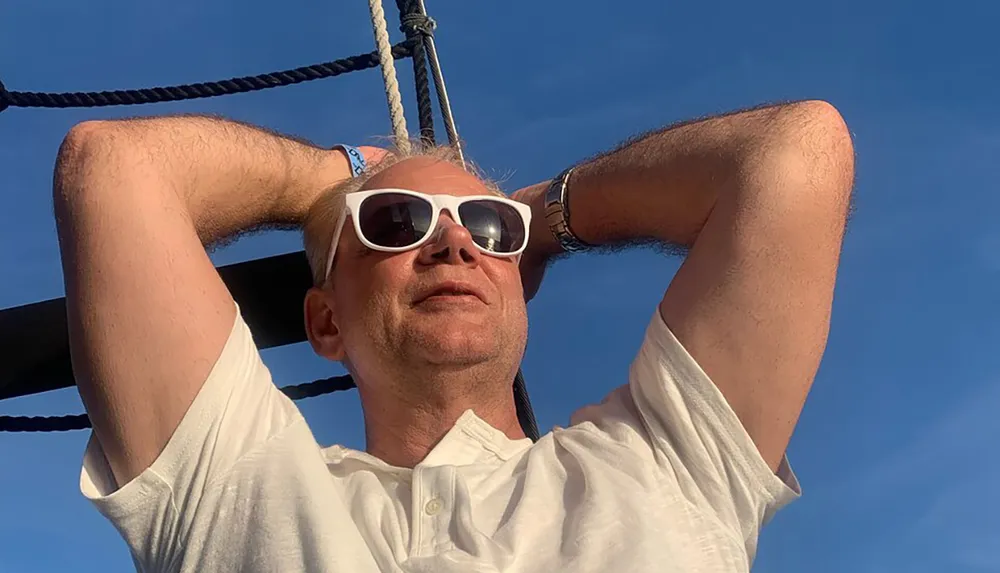 A man is relaxing with his arms behind his head wearing sunglasses and a white shirt with evident ropes and the clear blue sky in the background suggesting he might be on a boat