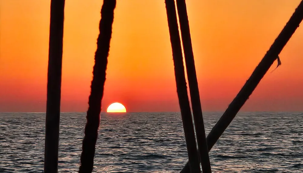 The image captures the serene beauty of a sunset with the sun dipping over the horizon of the sea framed by the silhouettes of ship rigging