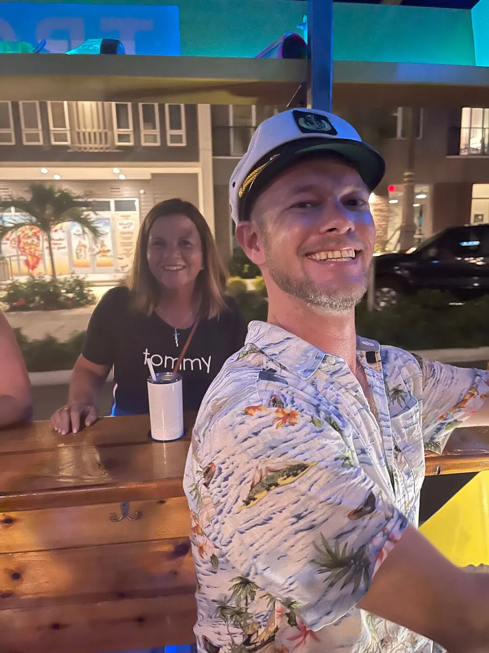 A man wearing a captains hat and a tropical shirt smiles for the camera at a nighttime social gathering while a woman stands in the background