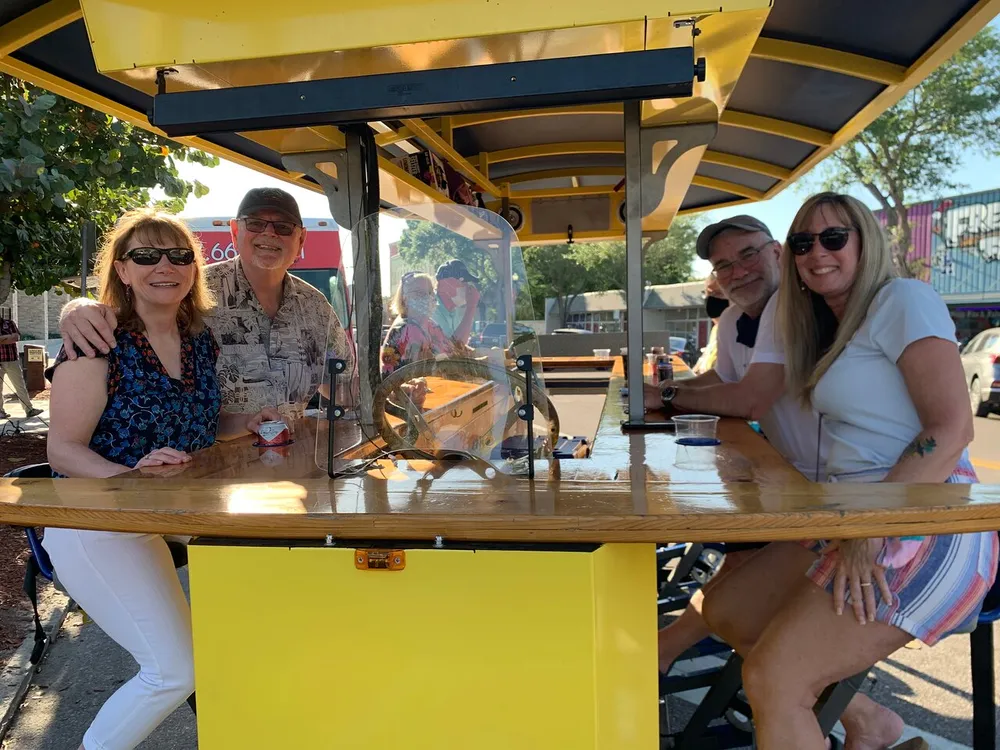 Four adults are smiling and sitting at a bar on wheels enjoying drinks on a sunny day