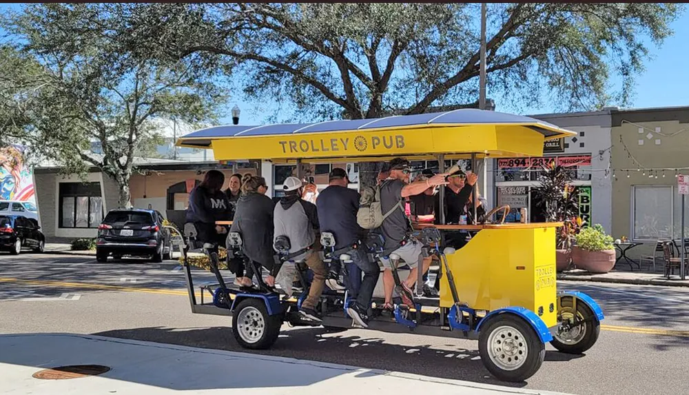 A group of people enjoys a ride on a pedal-powered Trolley Pub on a sunny day in an urban street setting
