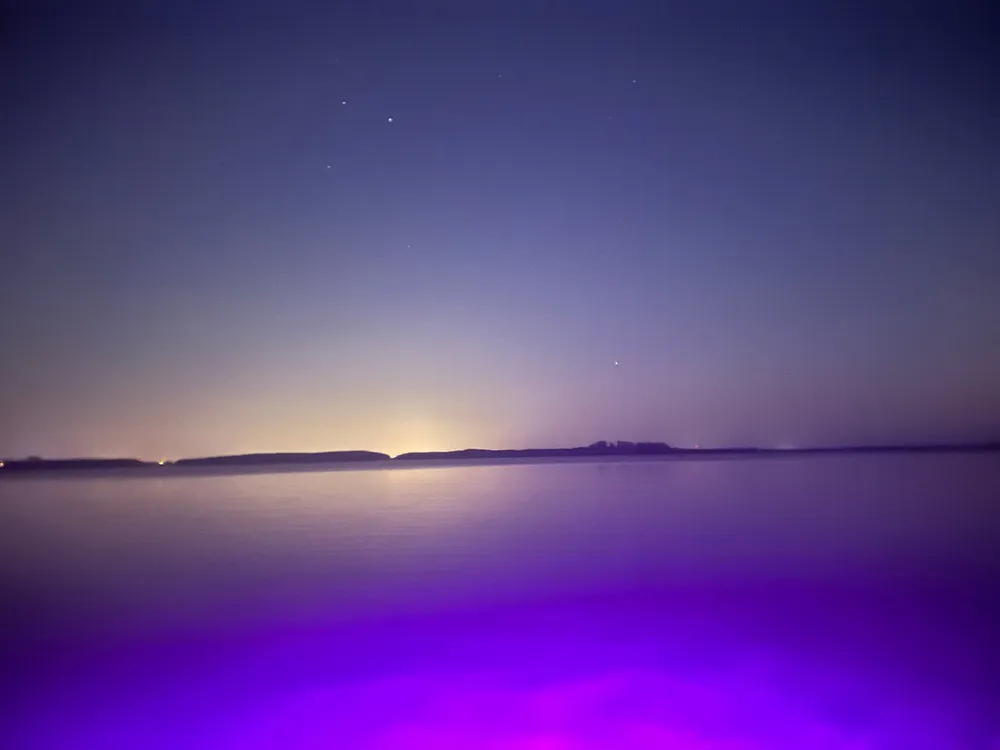The image captures a tranquil night scene with a vibrant purple foreground possibly water under a dusky sky with visible stars and a distant glow on the horizon