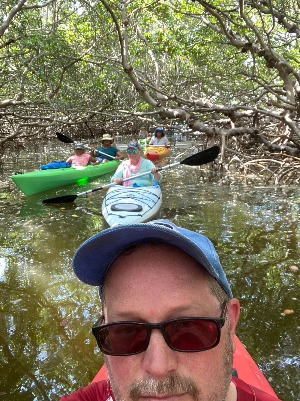 A group of people are kayaking in a mangrove forest with the forefront showing a selfie of a man wearing sunglasses and a baseball cap