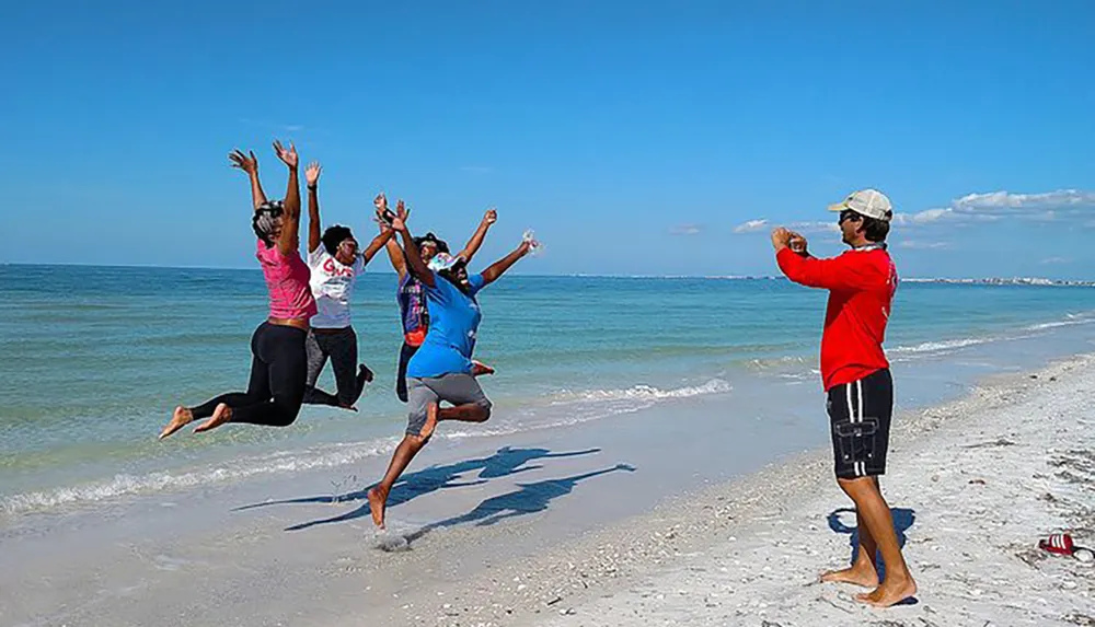 A group of people are joyfully jumping on a sunny beach while one person captures the moment with a camera