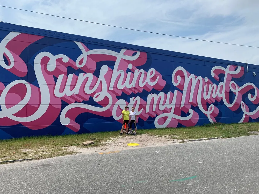 Two people stand by a vibrant blue wall with the phrase Sunshine on my Mind painted in large pink cursive letters