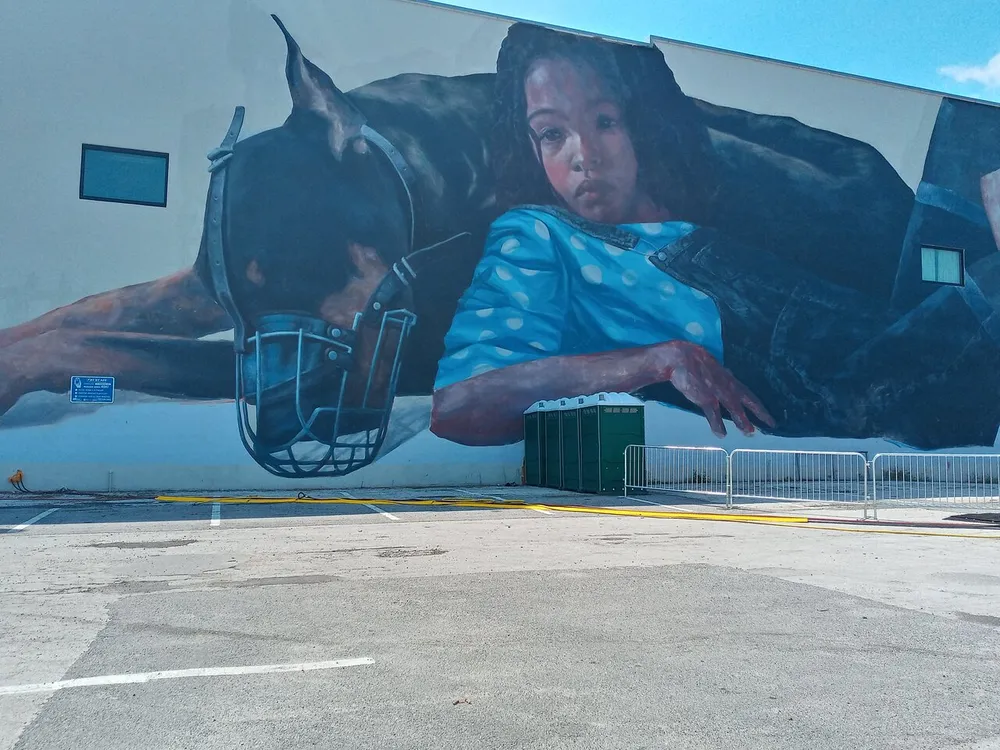 The image shows a large mural on the side of a building featuring a horse and a young girl contrasting with the mundane surroundings of a parking lot and portable toilets