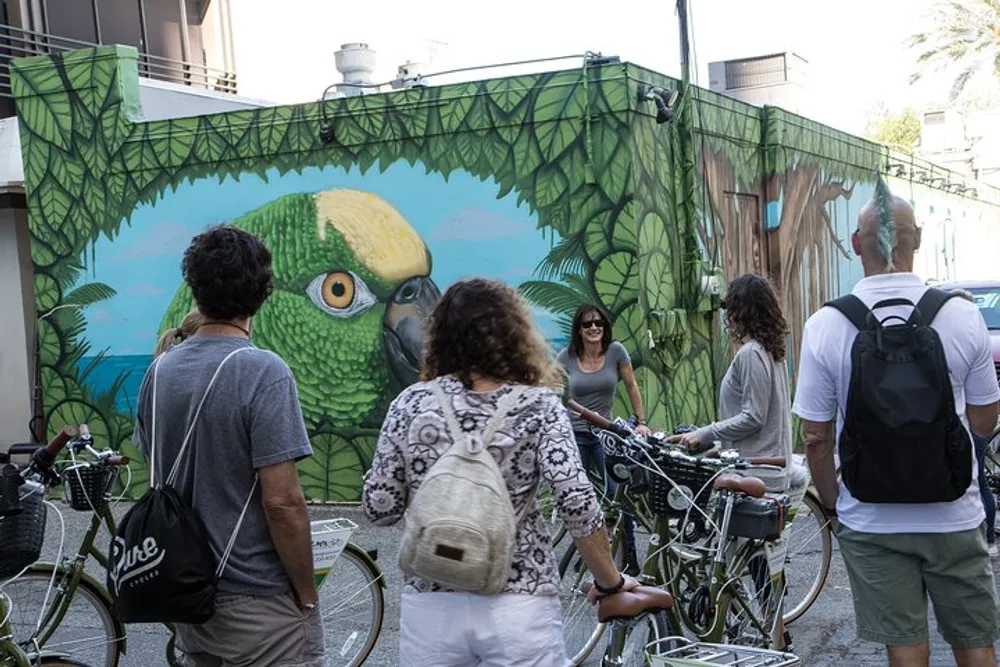 A group of people with bicycles are pausing to look at a vibrant mural of a parrot on an urban building