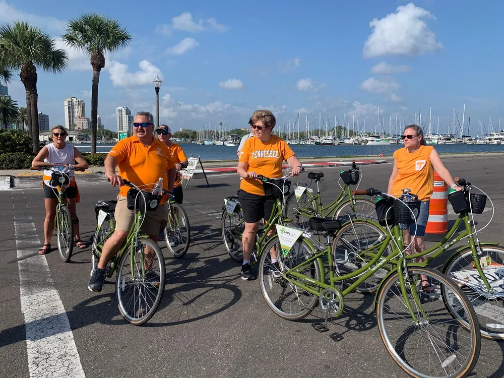 A group of people wearing orange shirts is ready to ride their rental bikes along a sunny waterfront promenade with palm trees and boats in the background