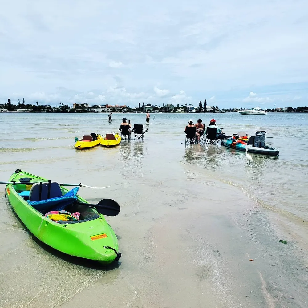 A group of people is enjoying sitting in chairs in shallow water near the shore with kayaks nearby indicating a leisurely day at the beach