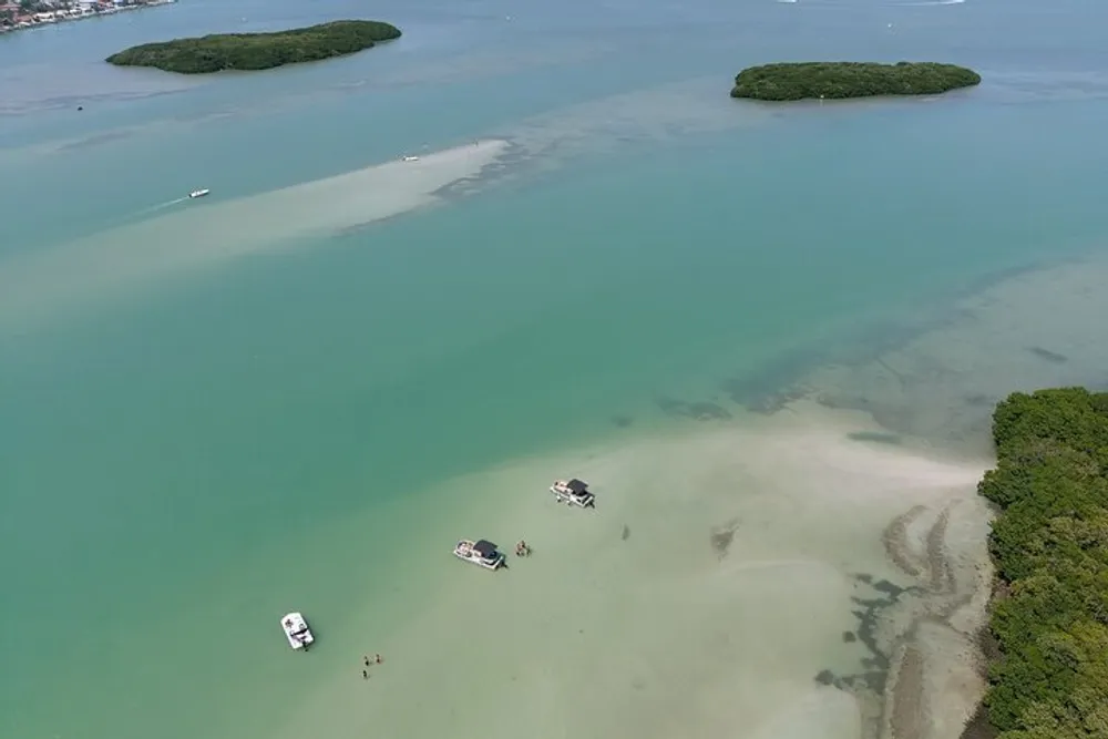 The image shows a serene aerial view of boats anchored near a sandbar with surrounding clear shallow water and patches of greenery where a few people appear to be enjoying a sunny day out on the water