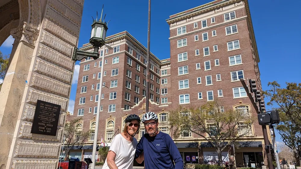 Two people wearing bicycle helmets are posing for a photo in a sunny urban setting flanked by historic-looking buildings and a decorated lamp post