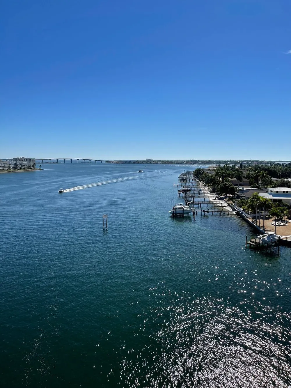 The image shows a sunny coastal scene with a clear blue sky a calm expanse of water with boat activity a long bridge in the distance and waterfront properties to the right