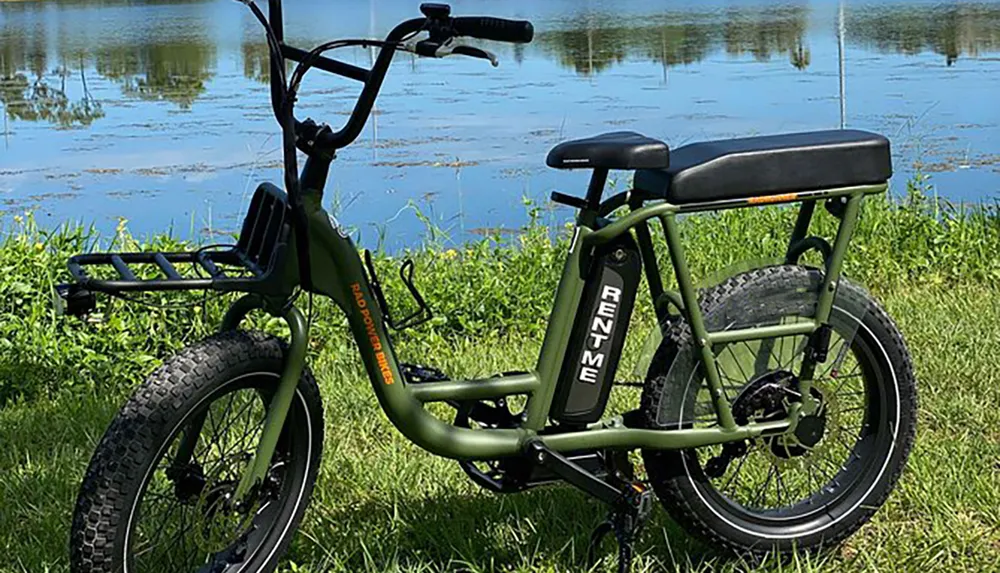 A sturdy green electric bike with thick tires is parked on grass by a lakeshore with the words RENT ME visible on its frame