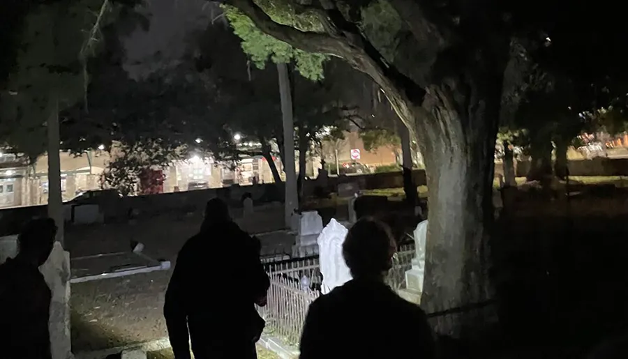 The image shows a group of people at night in what appears to be a cemetery with headstones and trees, illuminated by artificial light.