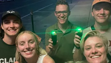 The image shows a group of five smiling people taking a group selfie at night, with two of them holding devices that emit green lights.