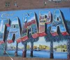 The image showcases a colorful mural on a wall with the words City of Tampa flanked by palm trees depicting iconic scenes and landmarks within each letter