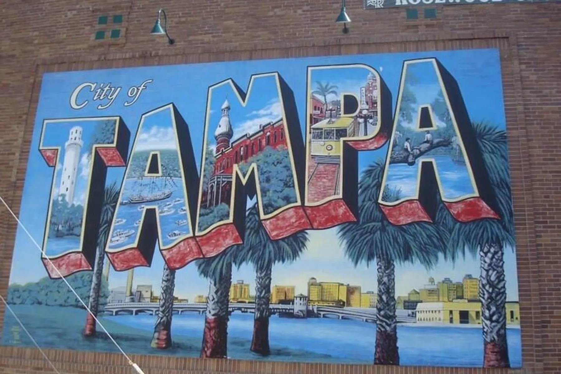 The image showcases a colorful mural on a wall with the words City of Tampa flanked by palm trees, depicting iconic scenes and landmarks within each letter.