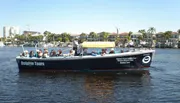 A group of people are on a dolphin-watching boat tour, enjoying a sunny day on the water with coastal buildings in the background.