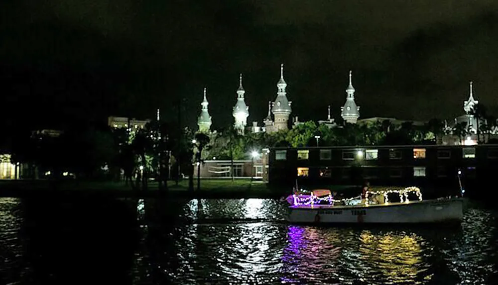 A boat adorned with lights floats on a body of water at night with a backdrop of a grand illuminated building with multiple spires