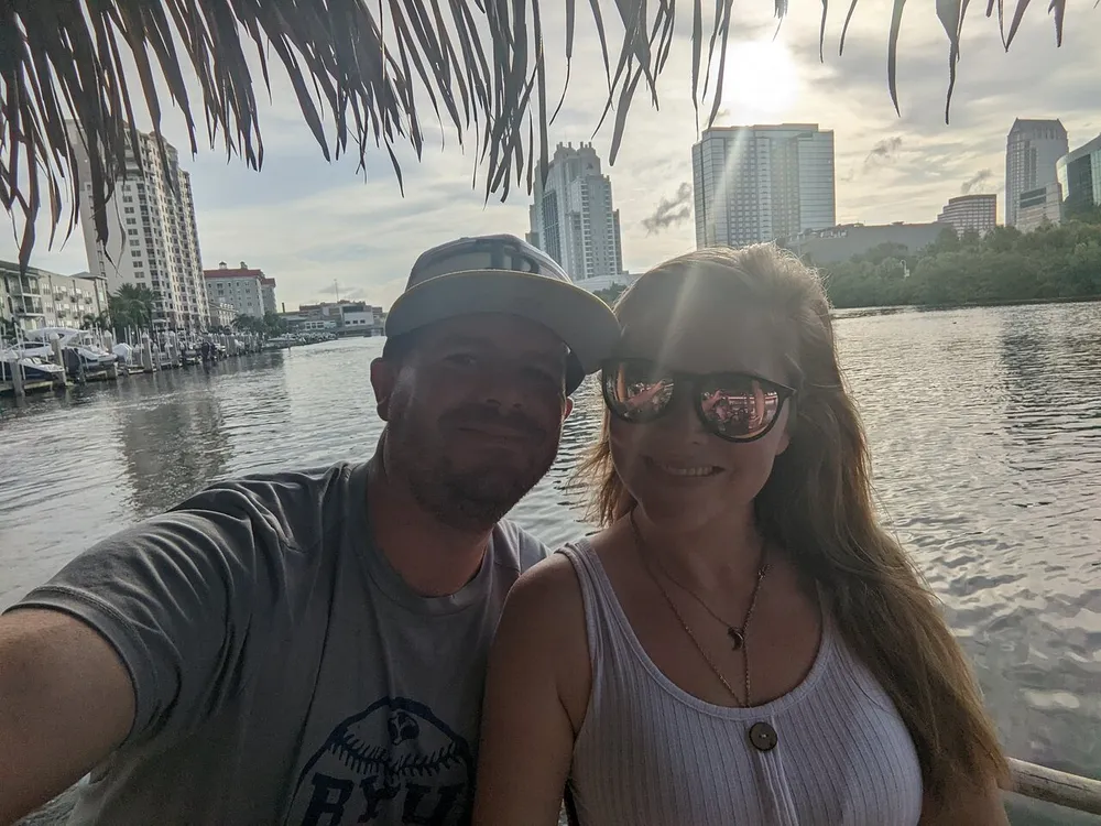 A man and a woman are posing for a selfie with a city skyline and waterfront in the background during what appears to be late afternoon or early evening