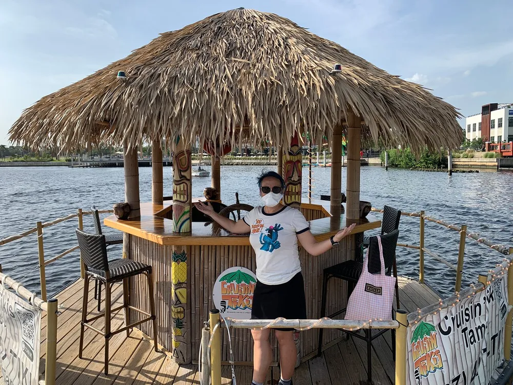 A person wearing a face mask stands beside a thatched-roof tiki bar which is apparently floating on the water with various signs and a city backdrop visible in the distance