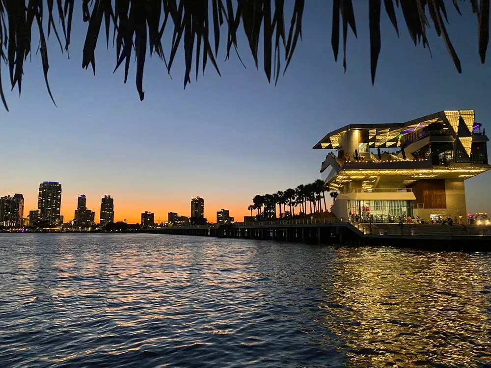 The image captures a modern waterfront building illuminated at twilight with a silhouette of palm trees and a city skyline against an orange-hued sky