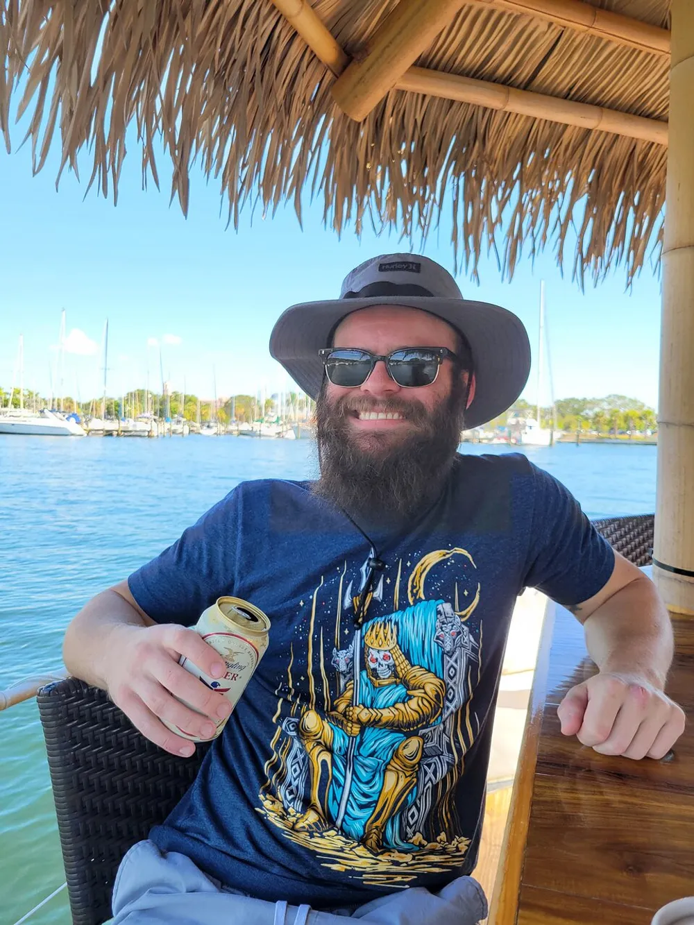 A smiling person wearing sunglasses a hat and a graphic t-shirt is sitting under a thatched roof with a drink overlooking a marina with boats