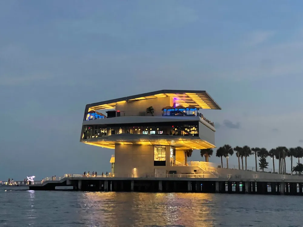 The image features a modern multi-level building extending over the water at twilight with illuminated interiors and people visible on the balconies against a backdrop of palm trees and a dusky sky
