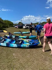 People prepare for a kayaking activity with several colorful kayaks laid out on the grass near parked vehicles under a sunny sky.