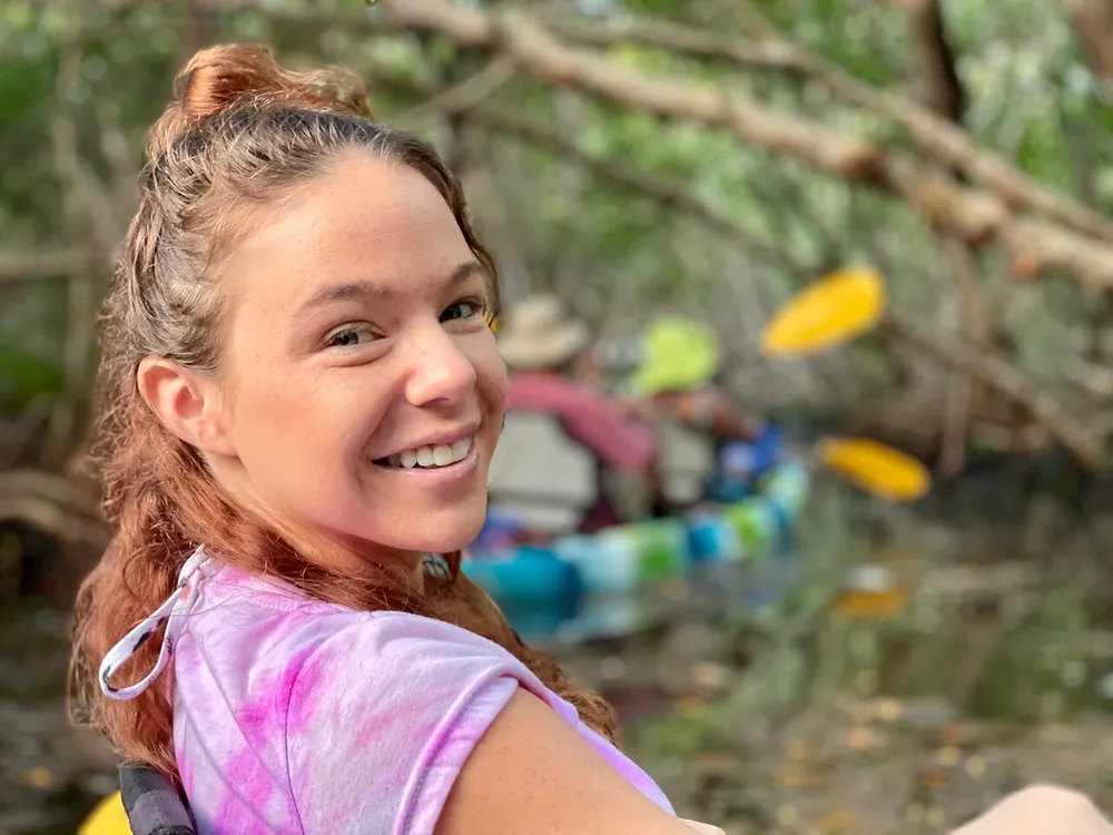A smiling person with their hair tied up is looking back at the camera with blurred kayakers in the background suggesting an outdoor recreational setting