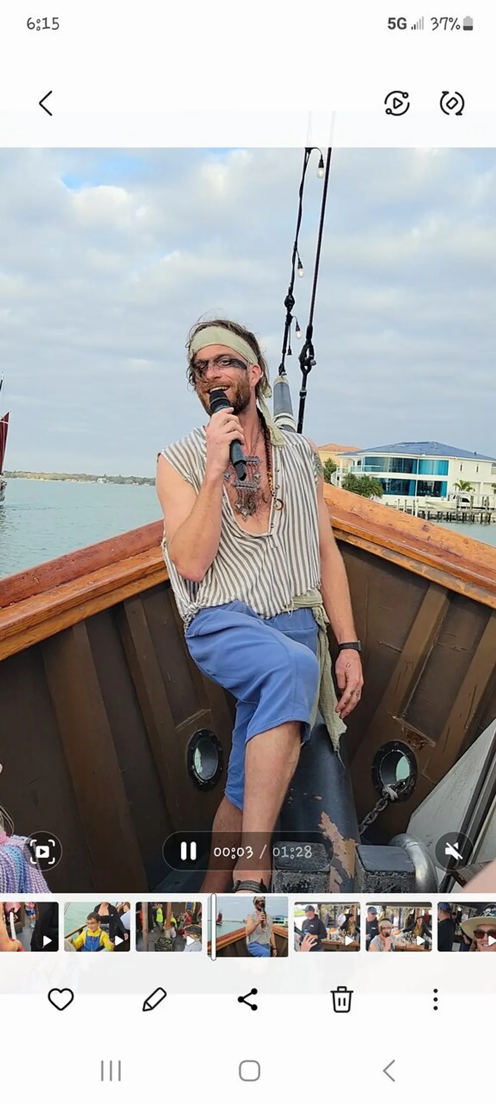 A person dressed as a pirate is sitting on the edge of a boat speaking into a microphone with a playful expression