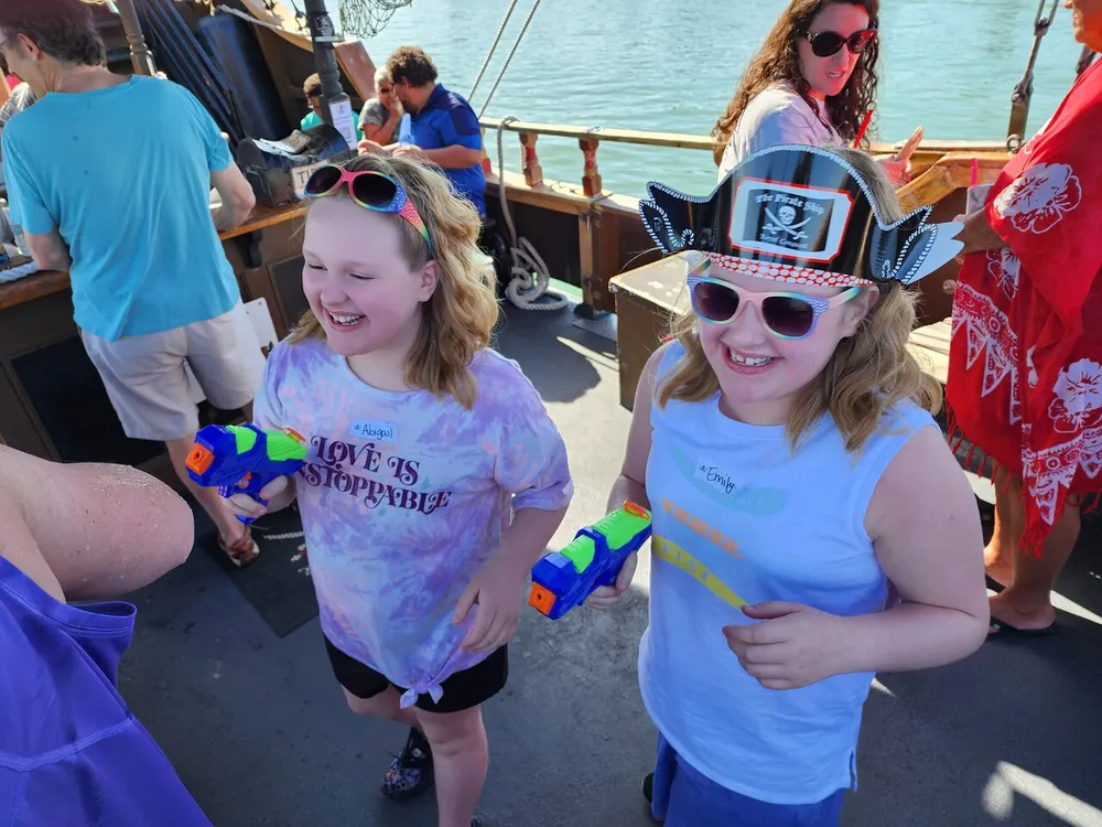 Two children are smiling joyfully one wearing a pirate hat while holding toy water guns on a sunny day aboard a boat with other passengers in the background