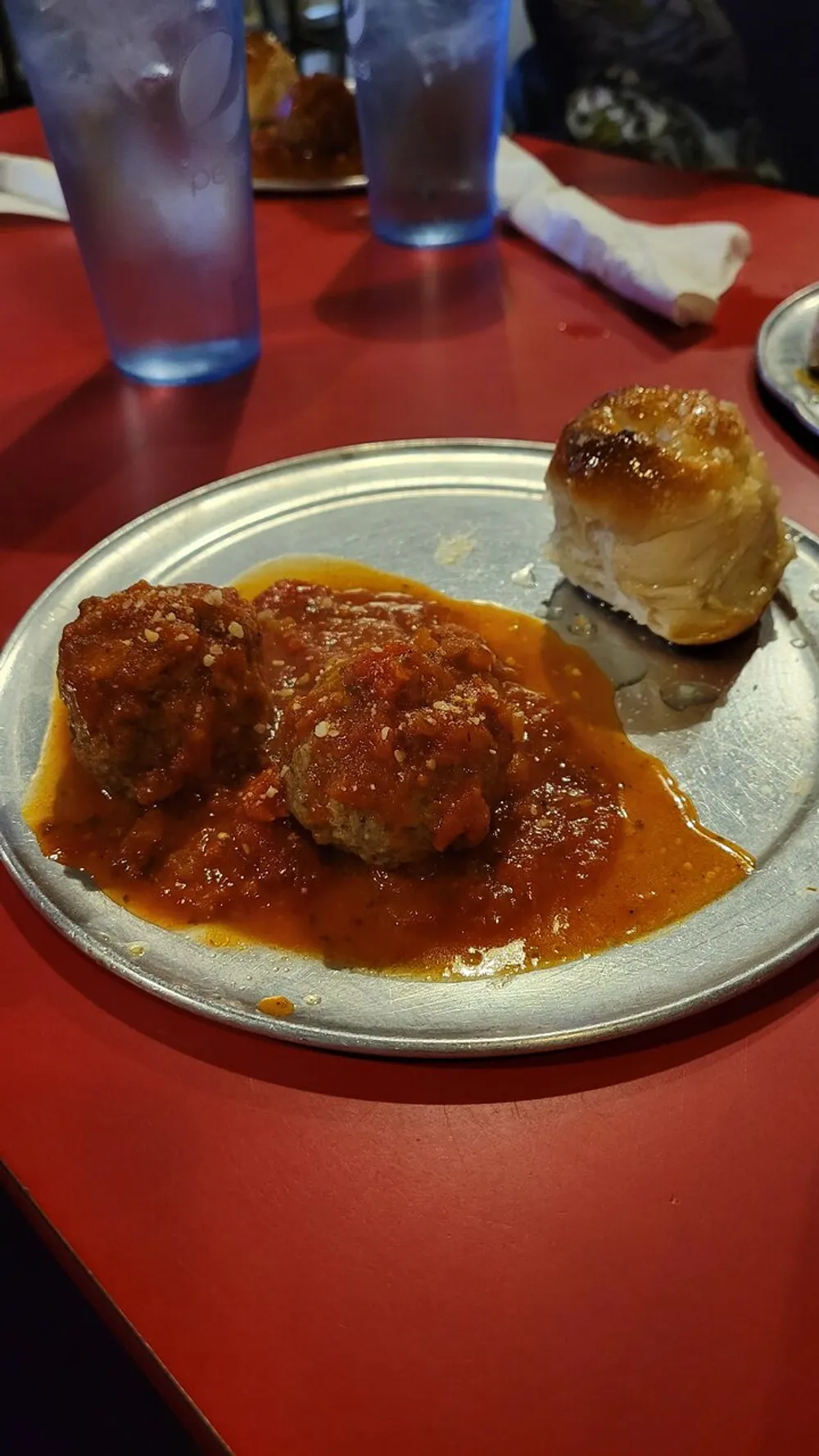 The image shows a plate with two meatballs covered in tomato sauce and sprinkled with grated cheese accompanied by a piece of bread on a table with a red surface and two glasses of water in the background