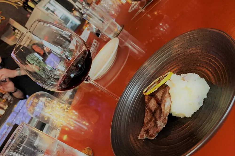 A glass of red wine is tilted perilously close to spilling while accompanying a plate with steak and mashed potatoes on a bar counter