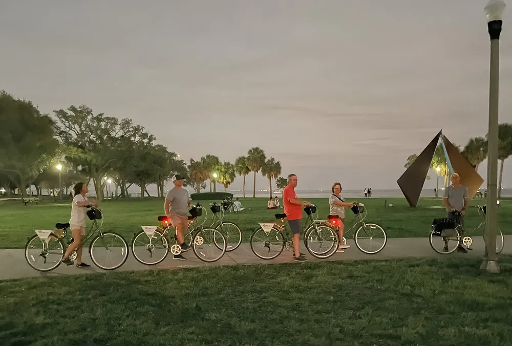 A group of people is standing with their bicycles in a park at dusk with some palm trees in the background and a sculpture on the right