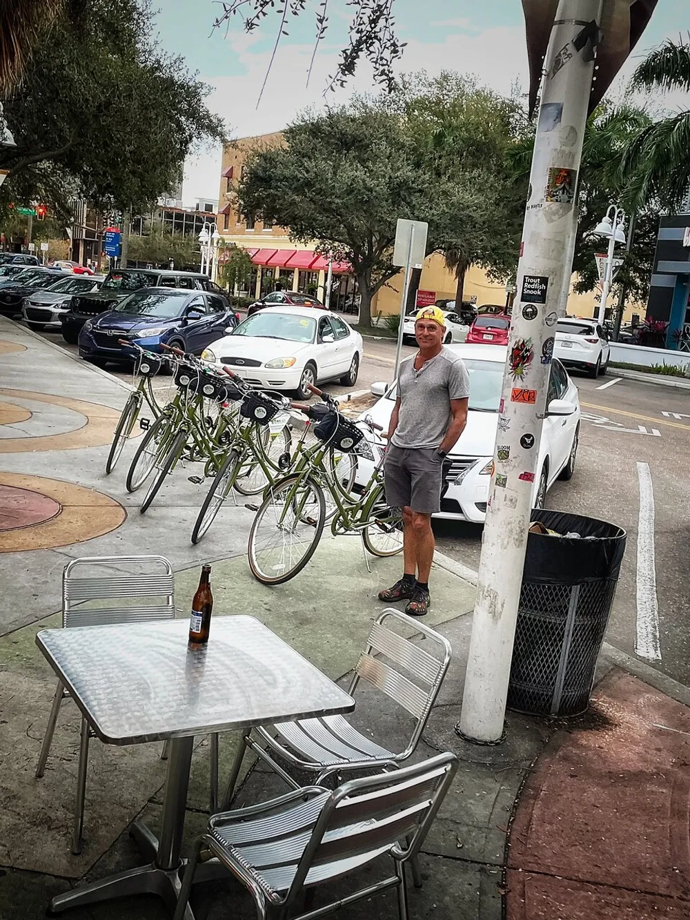 A person is standing on a sidewalk near a metal table with a bottle on it with parked bicycles and cars in the background in an urban setting