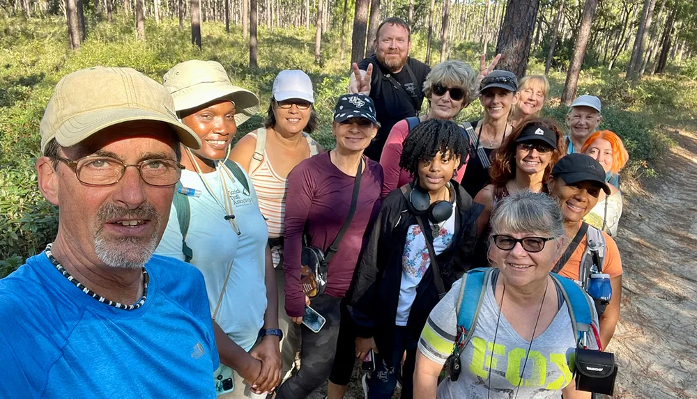 A diverse group of smiling people takes a selfie while enjoying a sunny day out on a hiking trail in a wooded area