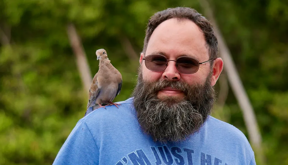 A smiling man with a beard is wearing sunglasses and a blue T-shirt while a dove perches on his shoulder