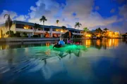 Two people are kayaking at dusk on a serene body of water with the colorful reflections of nearby buildings and ambient lights creating a picturesque scene.