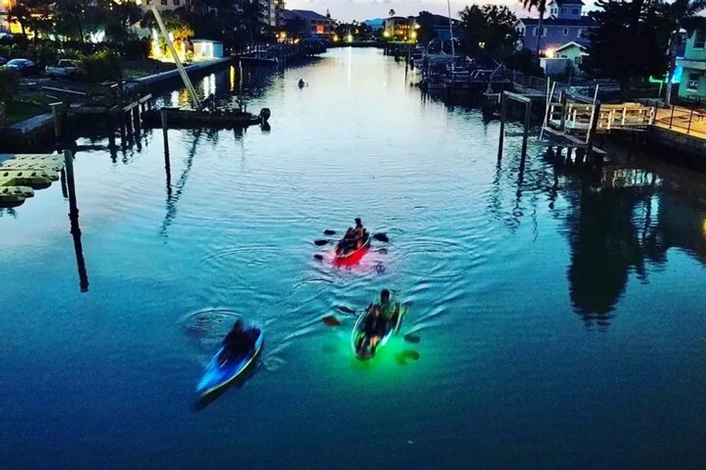 The image shows people kayaking at dusk on a tranquil waterway with the kayaks illuminated by colorful lights which create a striking reflection on the water