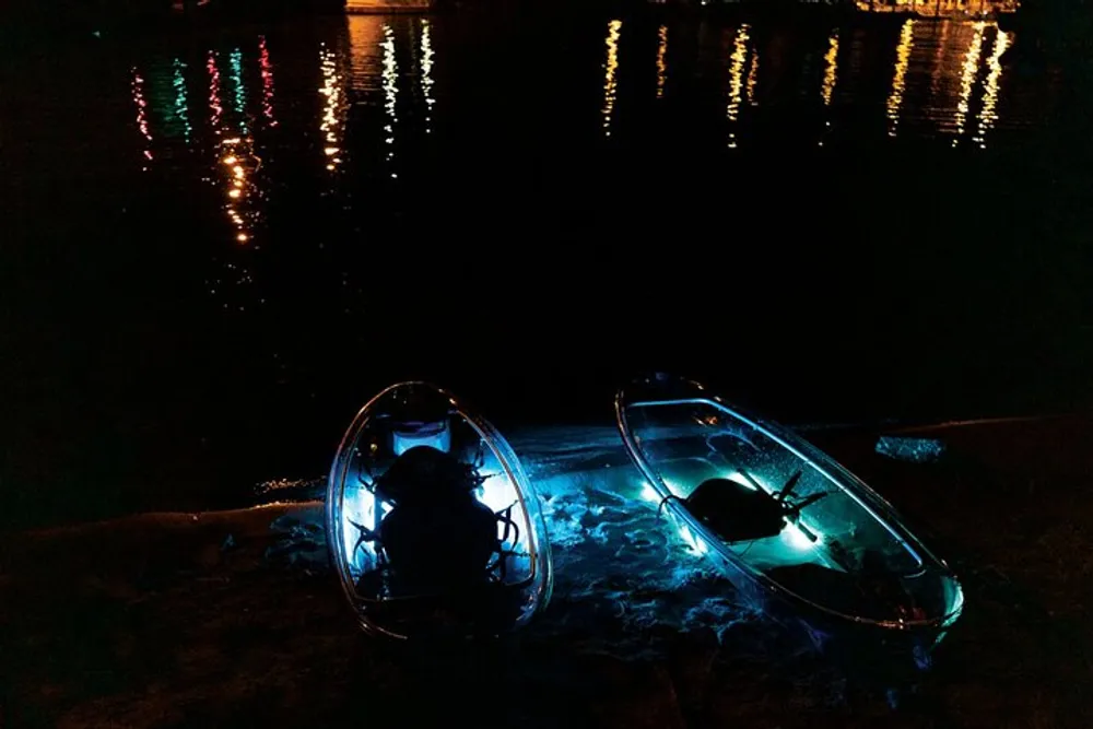 Two illuminated transparent kayaks rest on the water at night with colorful light reflections on the surface