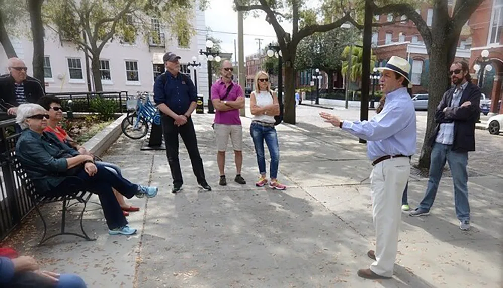 A man in a fedora hat is speaking to a group of attentive people outdoors possibly conducting a tour or giving a presentation