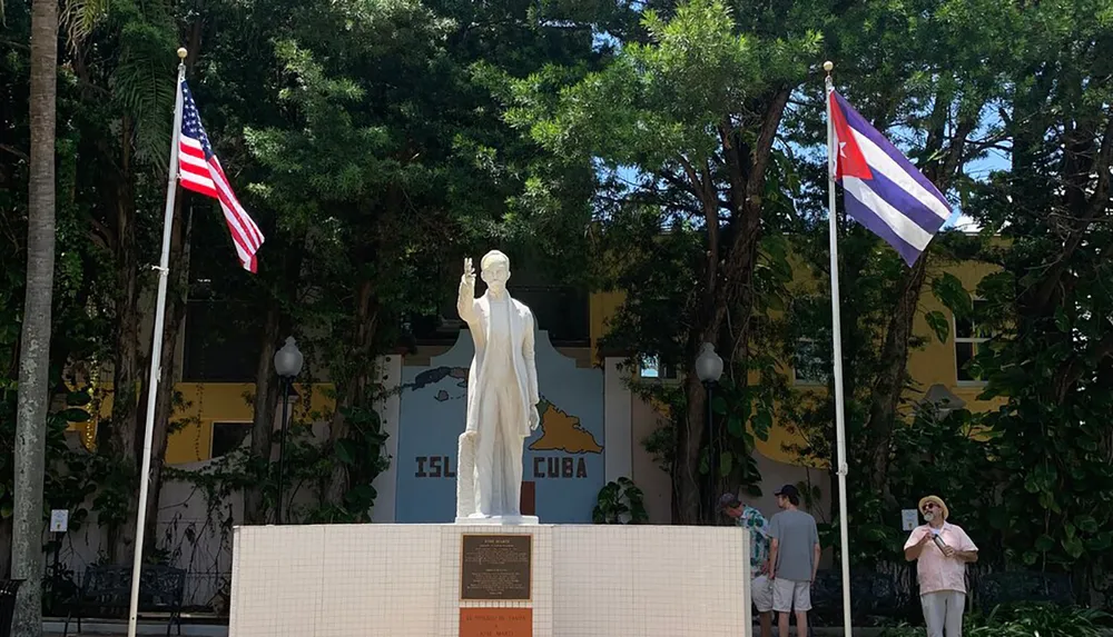 A statue is flanked by the flags of the United States and Cuba with visitors gathered around in a leafy outdoor setting