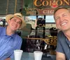 Two smiling men are taking a selfie in a caf with one wearing a straw hat and the other in a casual t-shirt and a Corona beer sign is visible in the background