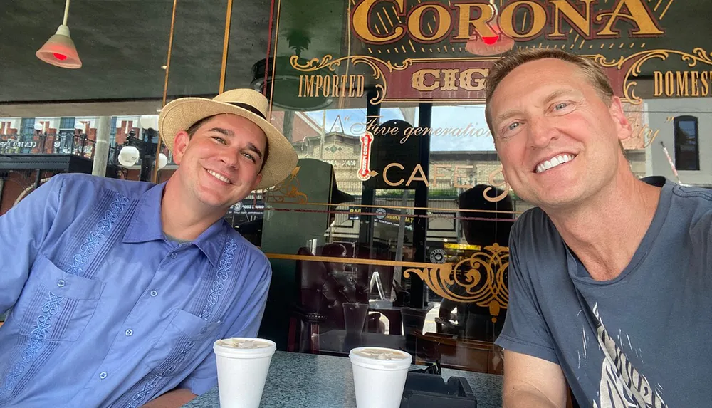 Two smiling men are taking a selfie in a caf with one wearing a straw hat and the other in a casual t-shirt and a Corona beer sign is visible in the background